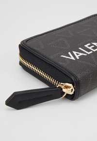 Portofel negru cu fermoar, fabricat din piele texturată, cu accesorii aurii la fermoar și un etichetă. Partea din față afișează "VALENTINO" în alb.