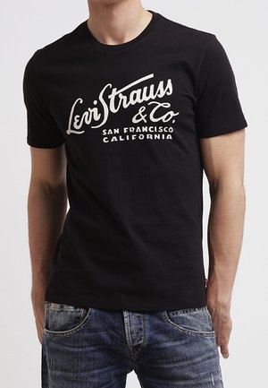 Homme portant un t-shirt noir Levi Strauss & Co. avec un logo texte blanc et un jean bleu, debout les bras détendus le long du corps.