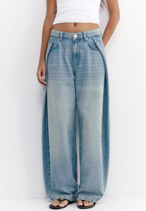 Frau trägt lockere, weit geschnittene, ausgebleichte blaue Jeans mit seitlichen Schlitzen, kombiniert mit einem weißen, kurz geschnittenen Tanktop und schwarzen Flip-Flops.