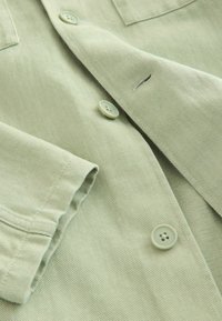 Tissu vert clair avec boutons et boutonnières visibles, montrant une texture détaillée et des coutures sur une chemise ou une veste.