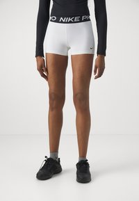 Bílé šortky Nike Pro s černým pasem, s designem loga. Ušité s černými sportovními botami a šedými ponožkami. Hladký povrch.