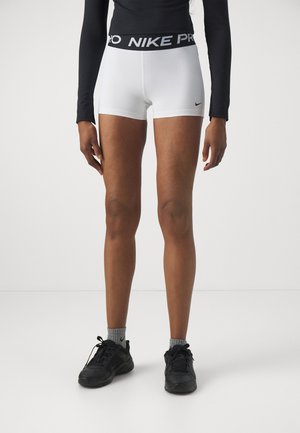 Shorts Nike Pro blancs avec une ceinture noire, arborant un design de logo. Portés avec des chaussures de sport noires et des chaussettes grises. Texture lisse.