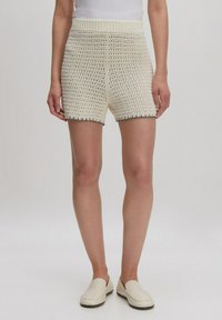 Gehaakte crèmekleurige shorts met een open weefpatroon, een geribbeld tailleband en zwarte detaleringen aan de onderkant. Gepaard met lichte, instap schoenen.