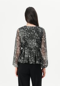 Blusa nera con motivo floreale, maniche lunghe trasparenti e vita arricciata. Presenta un delicato motivo floreale bianco su un tessuto leggero.