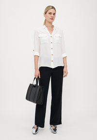 Witte button-up shirt met twee voorzakken, zwarte wijde broek, en een gestructureerde zwarte handtas, met glanzende zilveren schoenen.