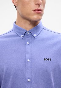 Svetlo modrá košeľa na gombíky s textúrovaným vzorom, so špicatým golierom a vyšitým logom "BOSS" v čiernej farbe.
