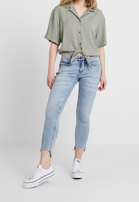 Camicia verde chiaro a maniche corte con nodo frontale, abbinata a jeans skinny azzurri con orlo sfrangiato e sneakers bianche basse.