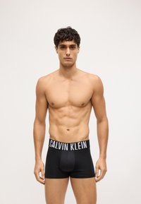 Boxeri scurți din bumbac negru, cu o centură albă lată pe care este inscripționat logo-ul "CALVIN KLEIN", croială standard și textură netedă. Modelul stă fără tricou.