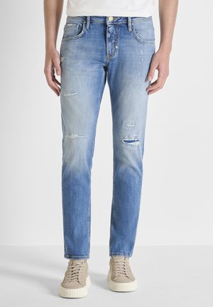 Homme portant un jean slim délavé bleu clair et des baskets beige, debout devant un fond blanc uni.