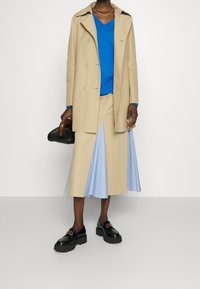 Trench-coat beige croisé porté sur un pull bleu à col en V, accompagné d'une jupe plissée bleu clair à fines rayures et de mocassins noirs épais.