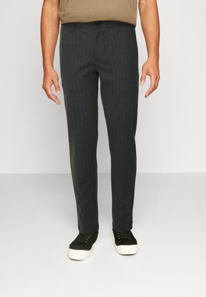 Hombre con pantalones ajustados de rayas grises oscuras, camisa marrón clara metida dentro y zapatillas negras de corte bajo con suelas blancas.