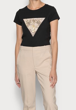 Woman wearing a black short-sleeve t-shirt with a leopard print Guess logo and beige high-waist trousers.