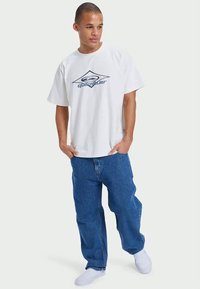 T-shirt branca de mangas curtas com o logótipo azul da Quiksilver, combinada com jeans de denim azul de corte largo. O modelo está em pé de forma descontraída com as mãos nos bolsos.