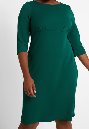 Ciemnozielona sukienka do kolan z rękawami do połowy przedramienia i dopasowaną talią na osobie o średnio-ciemnym odcieniu skóry.
