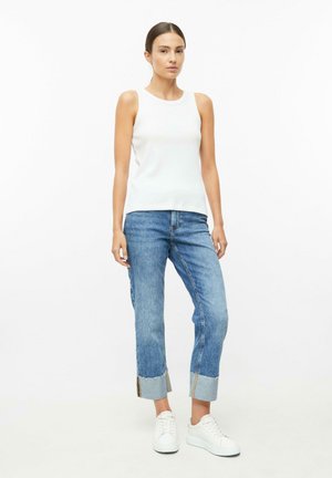 Vrouw draagt een mouwloos wit topje, cropped blauwe jeans met brede omslagen en witte sneakers, staand tegen een effen lichte achtergrond.