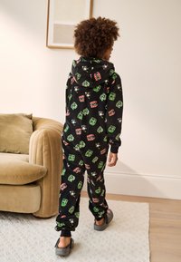 Black fleece onesie featuring colorful patterns of cartoon characters and shapes, with a hood and elastic cuffs, worn with gray slip-on shoes.