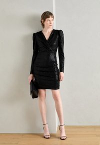 Robe à sequins noirs avec de longues manches ballons, décolleté en V, détails plissés et silhouette ajustée ; associée à une pochette noire et des talons à lanières.