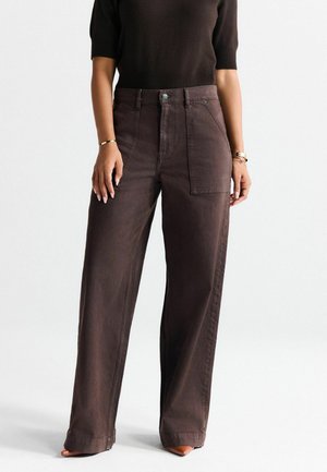 Woman wearing high-waist dark brown wide-leg pants with large front pockets, paired with a dark short-sleeve top and brown pointed shoes.