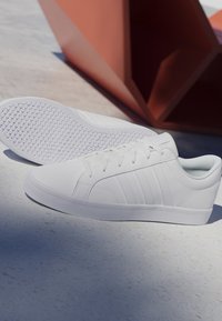 Par de zapatillas blancas bajas con suelas texturizadas colocadas sobre un suelo liso y claro cerca de una escultura geométrica marrón abstracta.