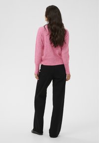 Rozā adīta džemperis ar garām piedurknēm un noapaļotu apakšmalu, kopā ar augstās jostas melnām biksēm un melnām plakano apavu kurpēm.