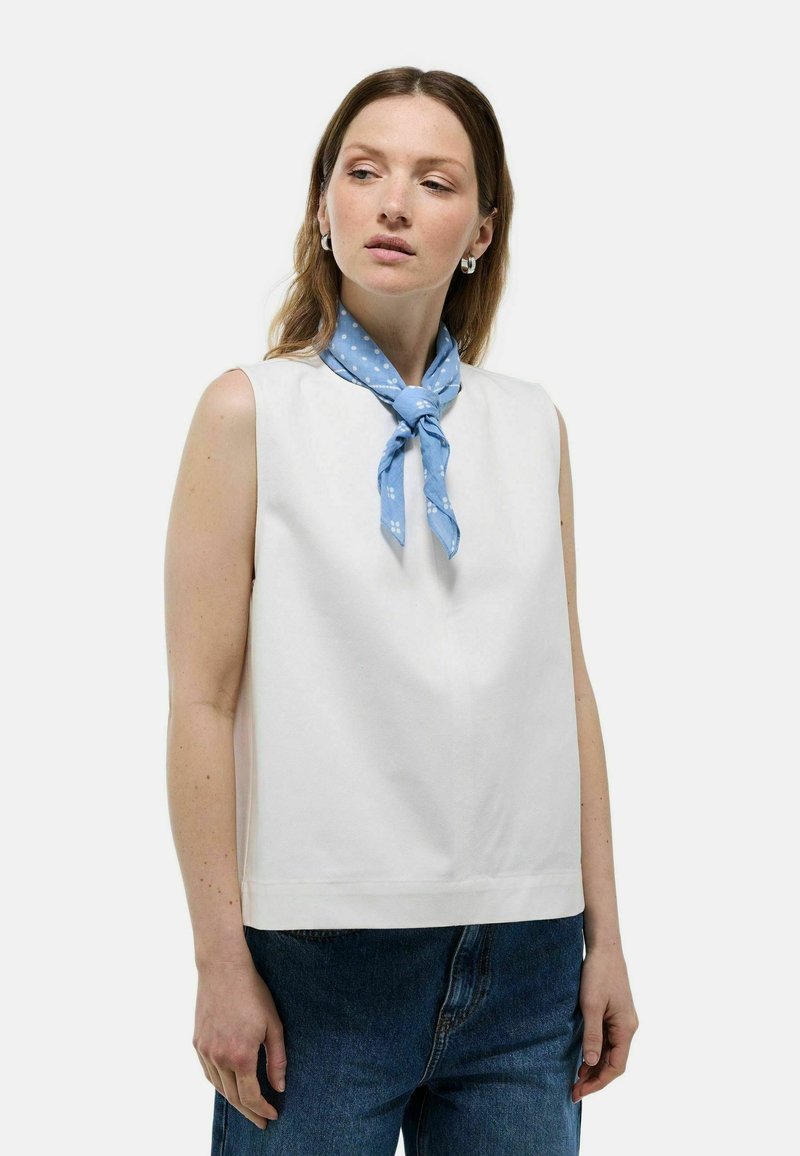 Top blanc sans manches avec un col montant et une coupe droite, associé à un foulard de cou à pois bleu clair. Tissu texturé avec des lignes épurées.