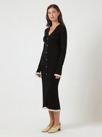 Γυναίκα που φοράει μαύρο midi φόρεμα με μακριά μανίκια και κουμπιά, με κρεμ βολάν στα μανίκια και το τελείωμα, και μπεζ χαμηλοτάκουνα παπούτσια τύπου slingback.