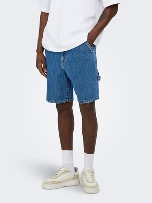 Mand iført mellem lange blå denimshorts, hvide crew sokker, beige sneakers og en hvid kortærmet skjorte, står med den ene hånd i lommen.