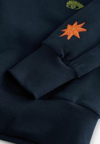 Felpa blu navy con polsini a coste, caratterizzata da un intricato disegno di un occhio verde e un ricamo a stella arancione sulla manica.