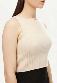 Beige ribbad crop-top med rundad halsringning och breda axelremmar; gjord av mjukt, strukturerat tyg och har en något fransig nederkant.