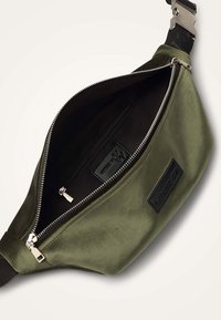 Banane en tissu vert olive à texture lisse, intérieur noir, fermeture éclair argentée et poche intérieure. Comprend une étiquette en cuir de marque.