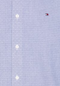 Světle modrá košile s malými bílými tečkami, bílé knoflíky vpředu a malé obdélníkové logo v červené, bílé a modré na hrudi.