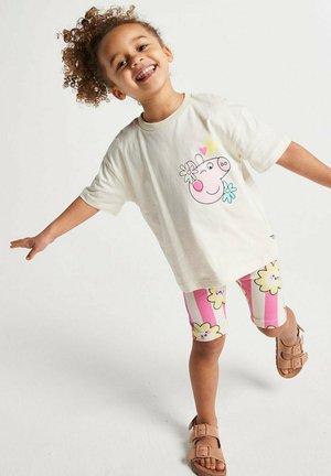Lächendes Kind, das auf einem Bein balanciert und ein weißes T-Shirt mit einem Cartoon-Schwein sowie gestreifte Shorts mit gelben Blumen und pinke Sandalen trägt.