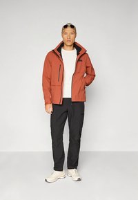 Casaco com fecho de correr, cor ferrugem, com capuz e detalhes pretos, combinado com calças pretas e ténis claros, contra um fundo simples.