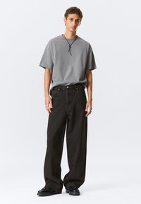 Mladý muž v volném šedém tričku, širokých černých džínách, černých botách a černém šňůrovém náhrdelníku, stojí před čistým bílým pozadím.