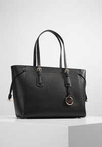 Bolso tote de piel negra de Michael Kors con doble asa, charm de logo dorado y correas ajustables con hebilla sobre una superficie de exhibición blanca.
