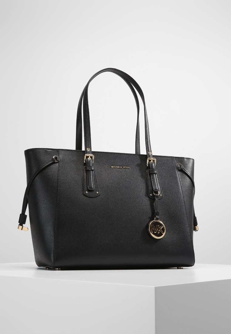 Bolso tote de piel negra de Michael Kors con doble asa, charm de logo dorado y correas ajustables con hebilla sobre una superficie de exhibición blanca.