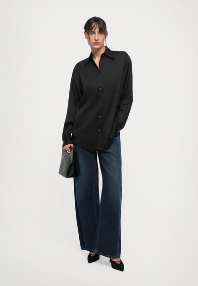 Schwarze Hemdbluse mit langen Ärmeln, kombiniert mit weitem, dunklem Jeans und schwarzen spitzen Pumps, hält eine kleine schwarze Clutch.