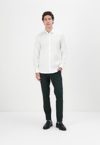 Weißes Langarmhemd mit geknöpftem Vorderteil, kombiniert mit dunkelgrüner Hose. Schwarze Schuhe runden das Outfit ab. Einfarbiger Hintergrund.