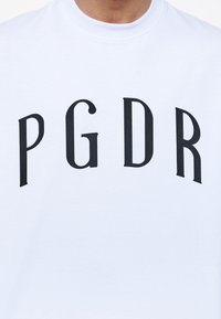 Biała bawełniana koszulka z dużym czarnym napisem graficznym "PGDR" w odważnej czcionce szeryfowej. Okrągły dekolt, krótkie rękawy, gładka tekstura.