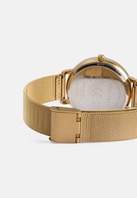 Orologio da polso in rete d'oro con cassa rotonda, fondo in acciaio inossidabile e chiusura a scatto. Design minimale, texture liscia e lucentezza metallica.