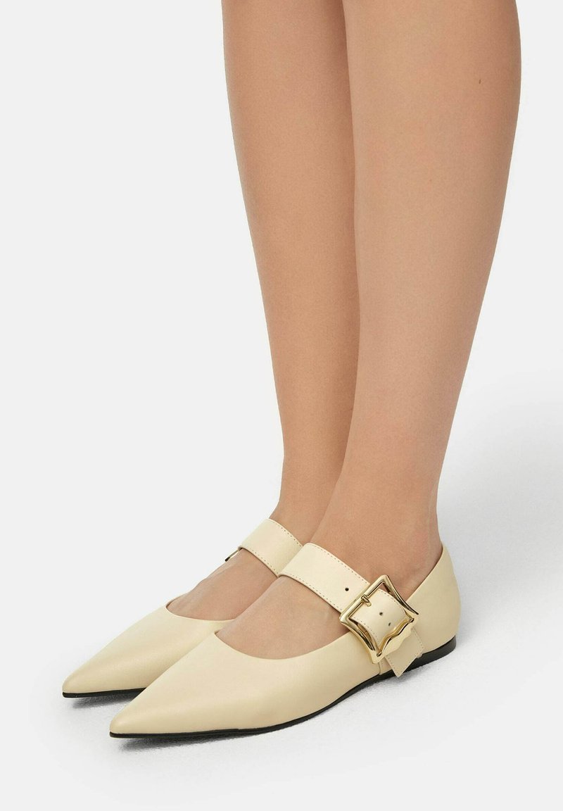 Ballerines en cuir crème avec un orteil pointu, une sangle sur le cou-de-pied ornée d'une boucle carrée dorée, et une semelle noire basse.