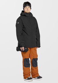 Zwarte buitenjas met capuchon, gecombineerd met oranje sneeuwbroeken met donkergrijze kniestukken. Model houdt zwarte handschoenen vast en draagt zwarte schoenen.