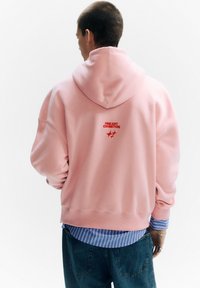 Rózsaszín kapucnis pulóver bő szabással, piros hímzett "FINE ART EXHIBITION" felirattal. Egy kék-fehér csíkos ingre van rétegezve.