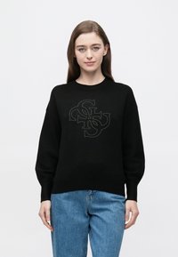 Melns džemperis ar apaļu kakla izgriezumu un garām piedurknēm, centrā ar izšūtu logotipa dizainu. Valkāts ar gaisziliem džinsiem.