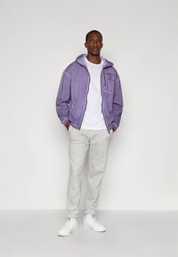 Μωβ φούτερ με φερμουάρ και μπροστινή τσέπη, λευκό t-shirt από μέσα, ανοιχτό γκρι παντελόνι φόρμας και λευκά αθλητικά παπούτσια. Λείο ύφασμα, casual εφαρμογή.