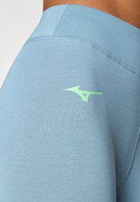 Lyseblå fritidssportbukser laget av glatt stoff. Har en grønn Mizuno-logo på benet og flatlock-sømmer langs sømmene.