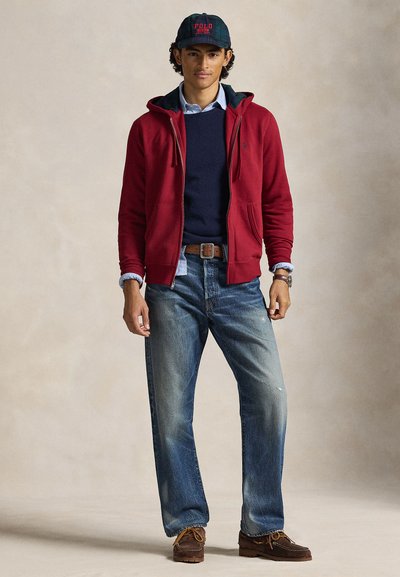 Camisola com fecho éclair vermelha sobre uma camisola azul-marinho, jeans azuis e sapatos estilo mocassim castanhos. O modelo usa um boné de padrão xadrez verde-escuro.