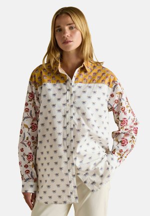 Woman with blonde hair wearing a buttoned, long-sleeve shirt with mixed floral patterns in white, yellow, and red tones, posing with one hand in pocket.