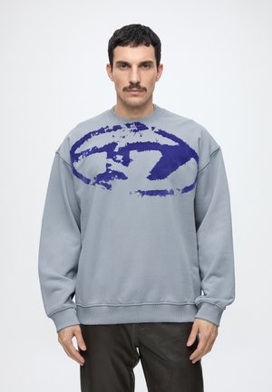 Uomo con capelli corti e scuri e baffi indossa una felpa grigio chiaro con un grande disegno astratto blu, in piedi contro uno sfondo chiaro e uniforme.