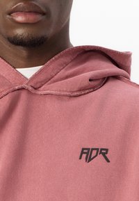Sweat-shirt à capuche en rose poussiéreux, fabriqué en tissu doux. Caractérisé par un logo noir "ADR" sur la poitrine et un col côtelé pour apporter de la texture.